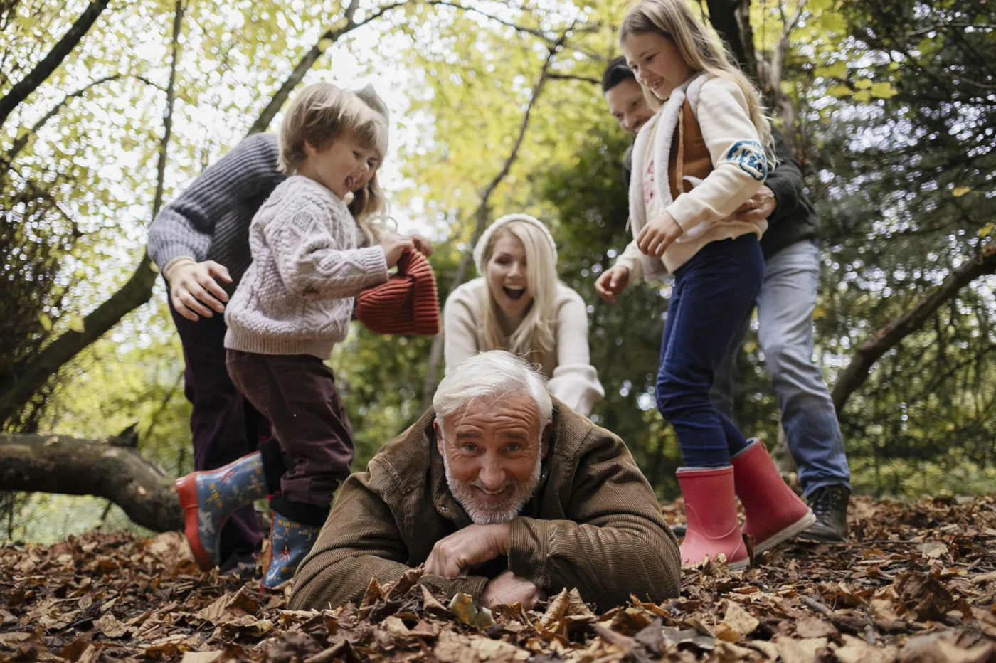 Familie spielt im Herbstwald, Kinder und Opa lachen im Laub am Boden.