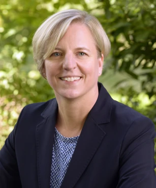 Porträt einer lächelnden Frau im Business-Outfit vor grünem, natürlichem Hintergrund.